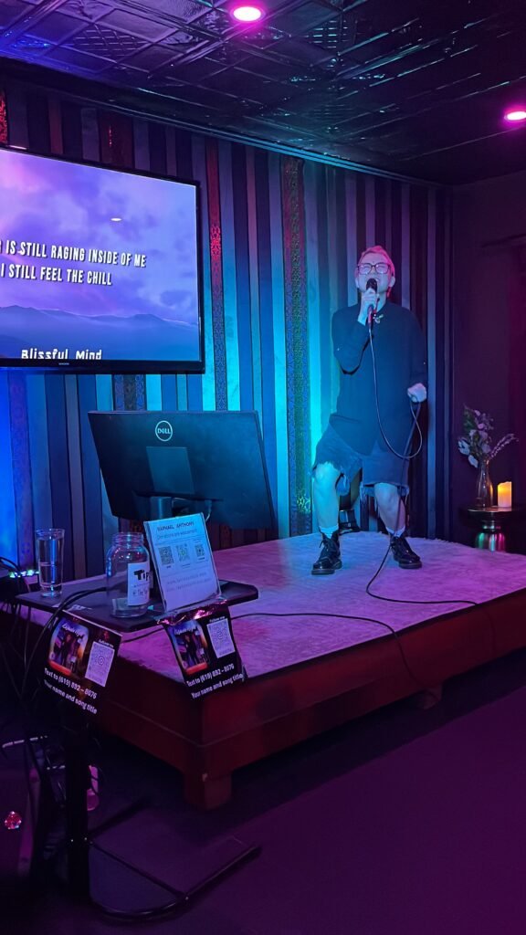 A young person with short pink hair, dressed in tall black boots and a black coat, is singing and emoting on a stage, in front of a TV screen with song lyrics, The mood is dark, with purple and blue light.