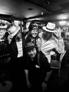 A black and white photograph of three men and a woman, dressed like the thirties, tipping their hats and smiling at the camera.