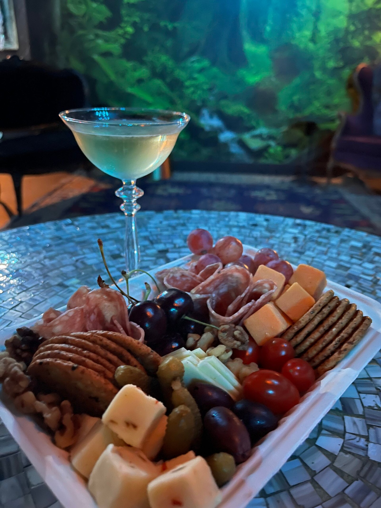 A glass of champagne next to a box of charcuterie, containing salami, cheese, grape tomatoes, crackers, cherries. In the background is the forest mural at The Wicked Wolf.